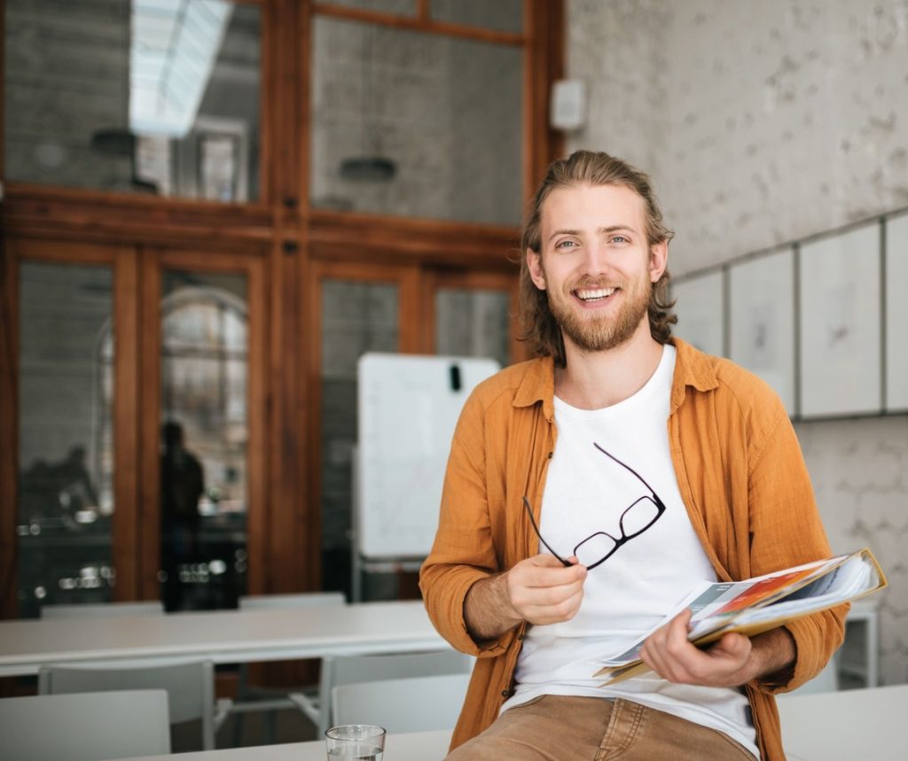 Junger Mann in kreativem Büro hält Unterlagen und lächelt in die Kamera. Modernes Arbeitsumfeld, Inspiration und positive Arbeitsatmosphäre.