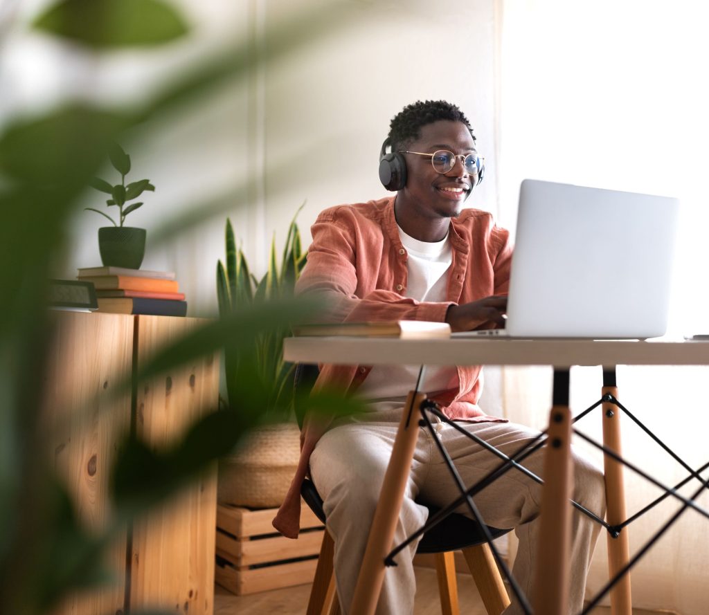 Ein junger Mann mit Brille und schwarzen Kopfhörern sitzt lächelnd an einem weißen Schreibtisch und arbeitet konzentriert an seinem Laptop.