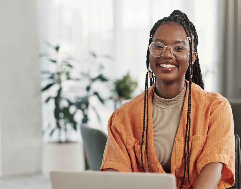 Eine junge Frau mit langen Braids und Brille sitzt lächelnd vor ihrem Laptop in einer hellen, modern eingerichteten Wohnung.