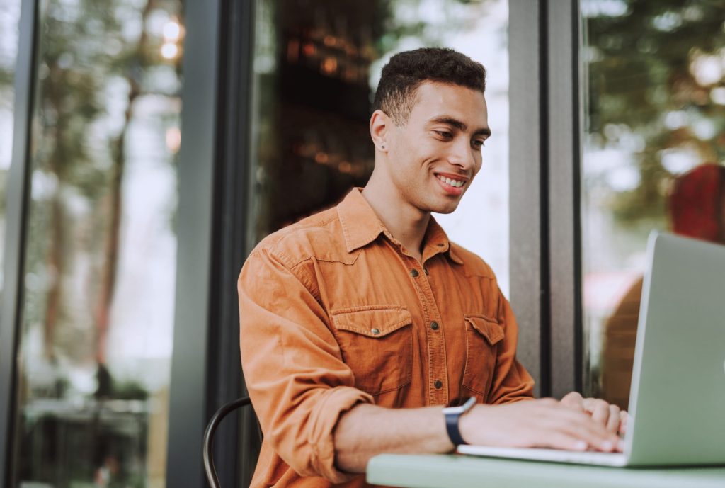 Junger Mann sitzt draußen im Café, arbeitet am Laptop und lächelt dabei.