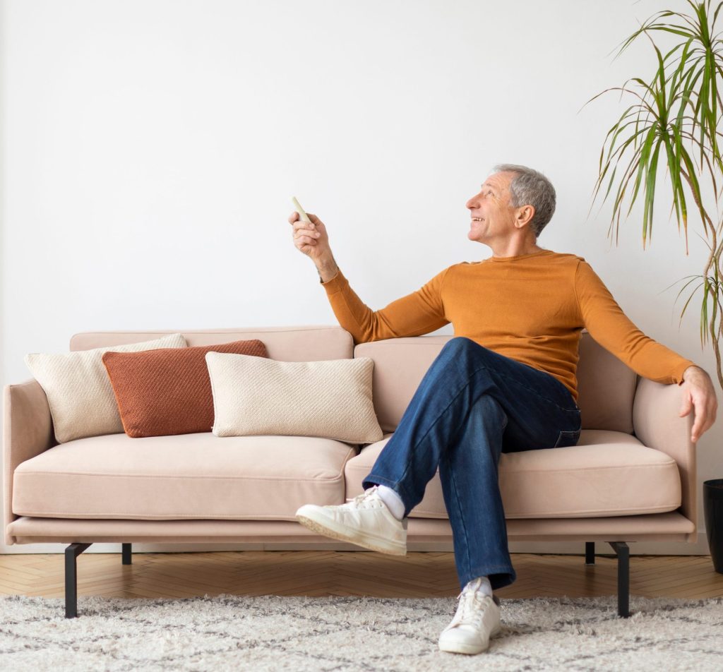 Ein lächelnder älterer Mann in einem orangefarbenen Pullover sitzt auf einem hellen Sofa und bedient eine Fernbedienung