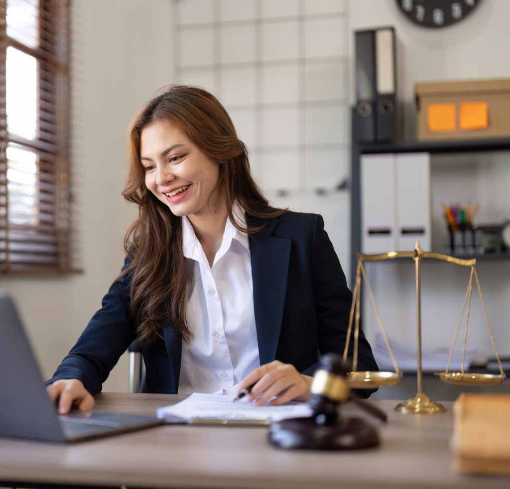 Eine lächelnde junge Anwältin im dunklen Business-Outfit arbeitet an ihrem Laptop in einer modernen Kanzlei