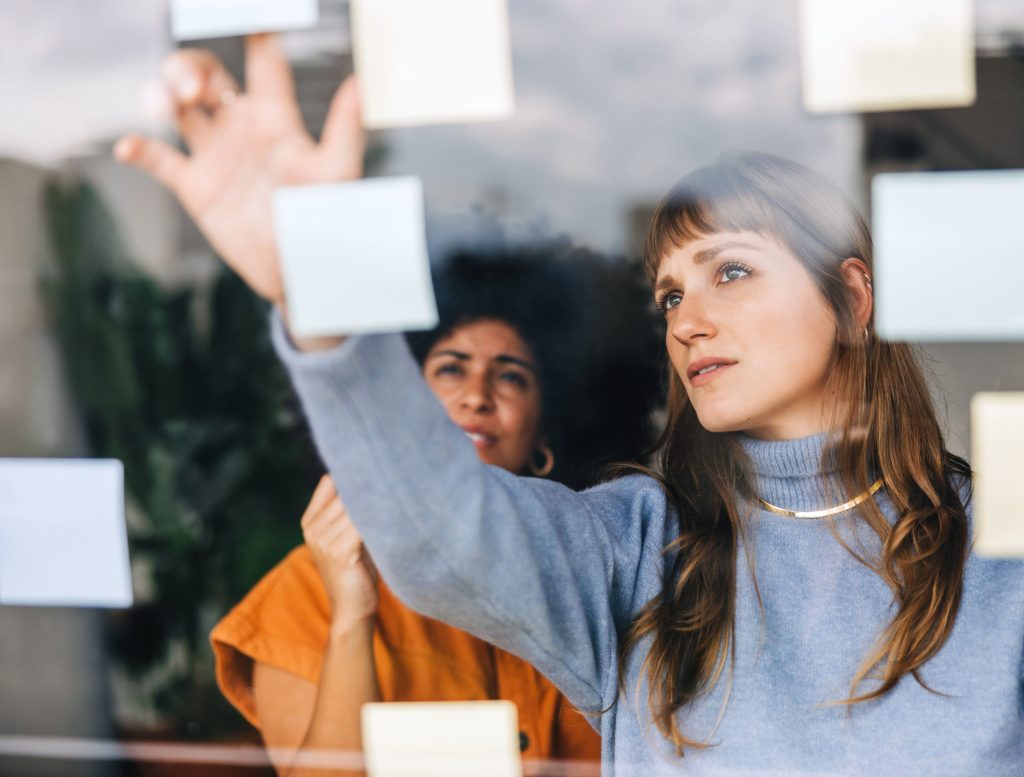 Zwei junge Frauen arbeiten konzentriert an einer Glaswand und ordnen gelbe sowie weiße Haftnotizen bei einem Brainstorming an