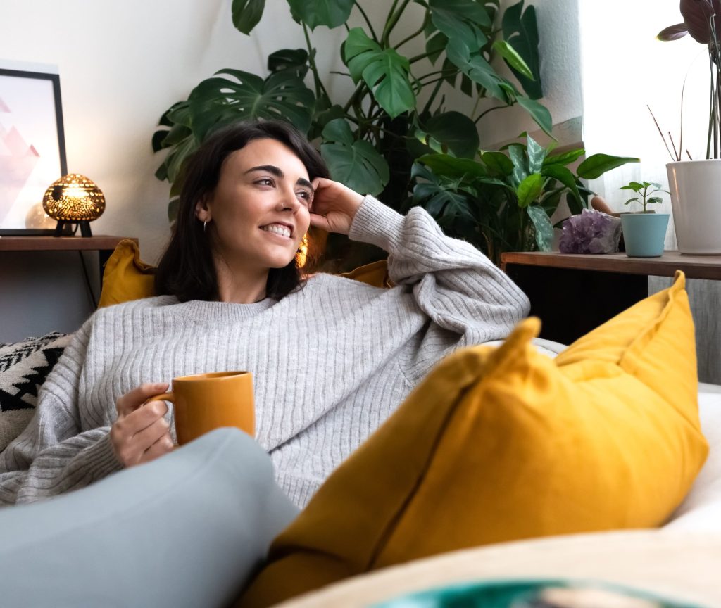 Eine lächelnde junge Frau entspannt sich in einem grauen Strickpullover auf einem hellen Sofa, hält eine gelbe Kaffeetasse und schaut gemütlich zur Seite