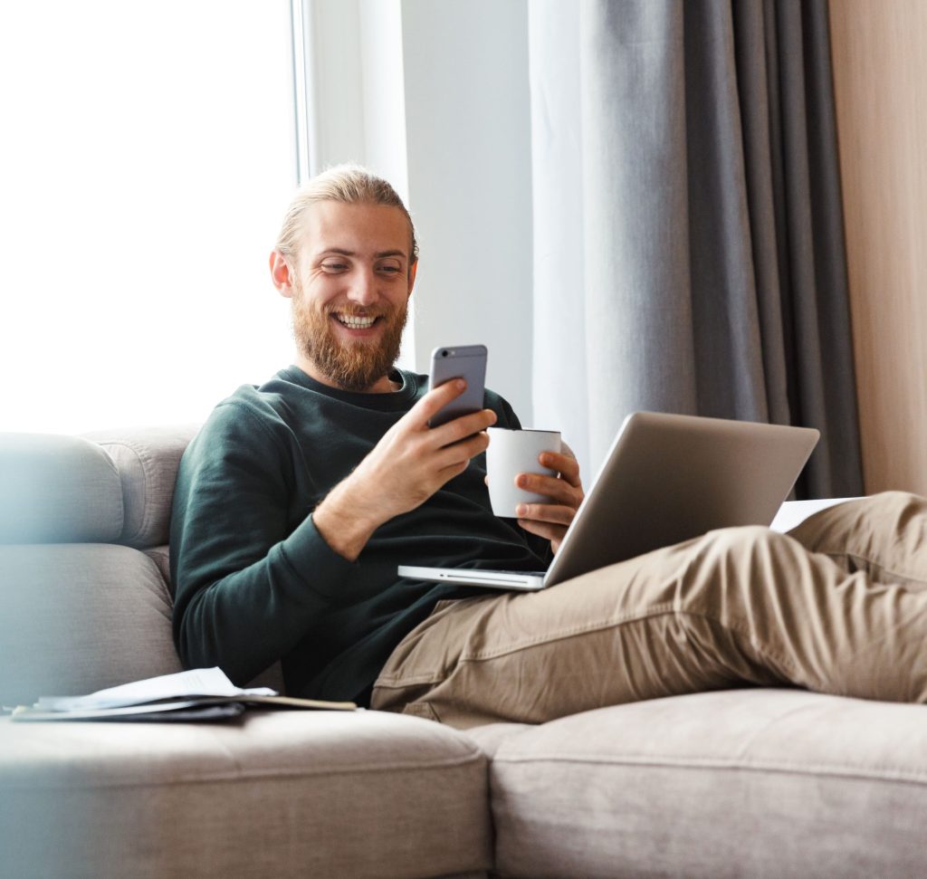 Ein lächelnder Mann mit Bart sitzt entspannt auf einem grauen Sofa, hält eine Kaffeetasse und schaut auf sein Smartphone, während ein Laptop auf seinem Schoß liegt