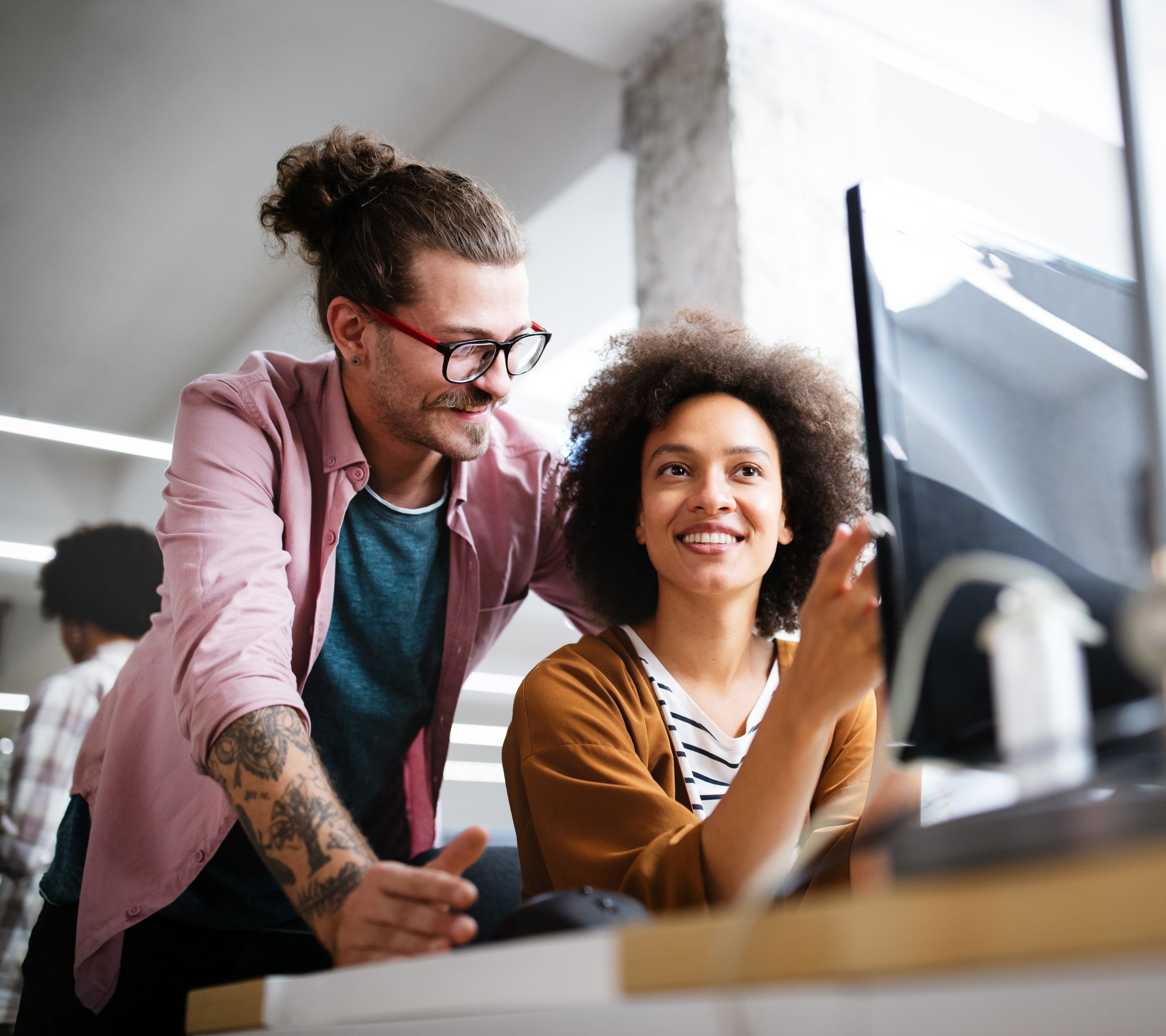 Eine junge Frau mit langen Braids und Brille sitzt lächelnd vor ihrem Laptop in einer hellen, modern eingerichteten Wohnung.