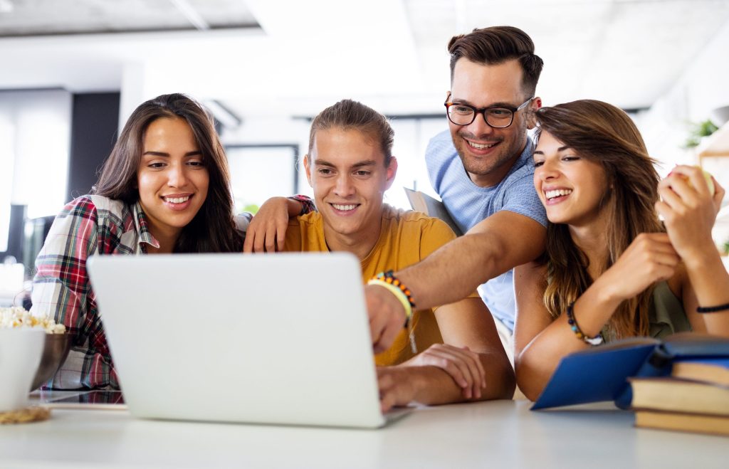 Vier junge, lachende Erwachsene sitzen gemeinsam an einem Tisch in einer modernen Küche und blicken gespannt auf den Bildschirm eines Laptops.