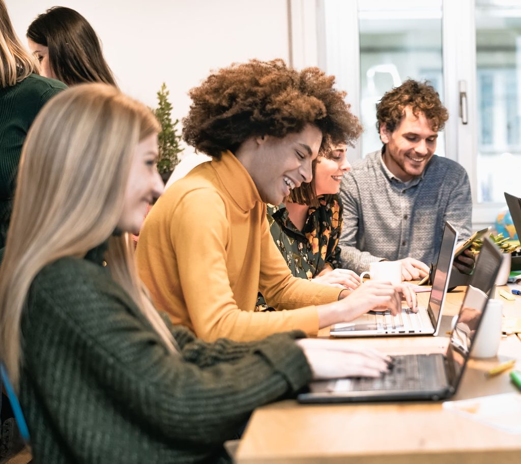 Eine Gruppe junger, lächelnder Menschen arbeitet gemeinsam an einem langen Holztisch mit Laptops und Tablets in einem hellen Coworking-Space