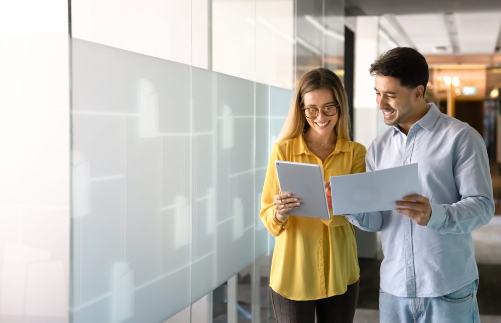 Eine junge Frau und ein Mann stehen lächelnd in einem modernen Büroflur und besprechen Dokumente sowie Inhalte auf einem Tablet