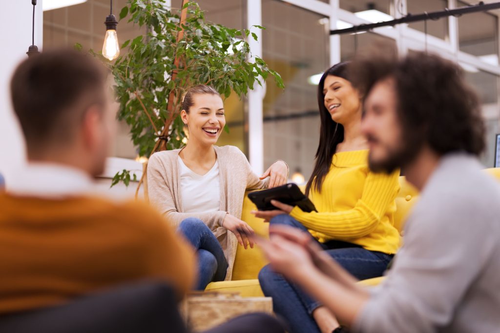 Vier junge Menschen sitzen entspannt in einem modern gestalteten Büro zusammen, lachen und tauschen sich über ein Tablet aus