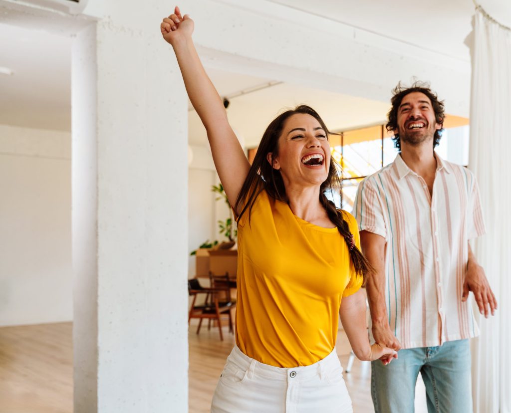 Ein junges Paar feiert ausgelassen in einer hellen, leeren Wohnung; die Frau im gelben T-Shirt reißt jubelnd einen Arm hoch, während der Mann im Hintergrund lacht.