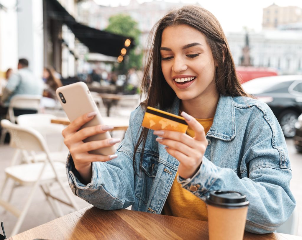 Eine lächelnde junge Frau in einer Jeansjacke sitzt an einem Holztisch im Freien, hält eine goldene Kreditkarte und nutzt ihr Smartphone. Vor ihr steht ein Kaffeebecher zum Mitnehmen in einer belebten Straßenszene.