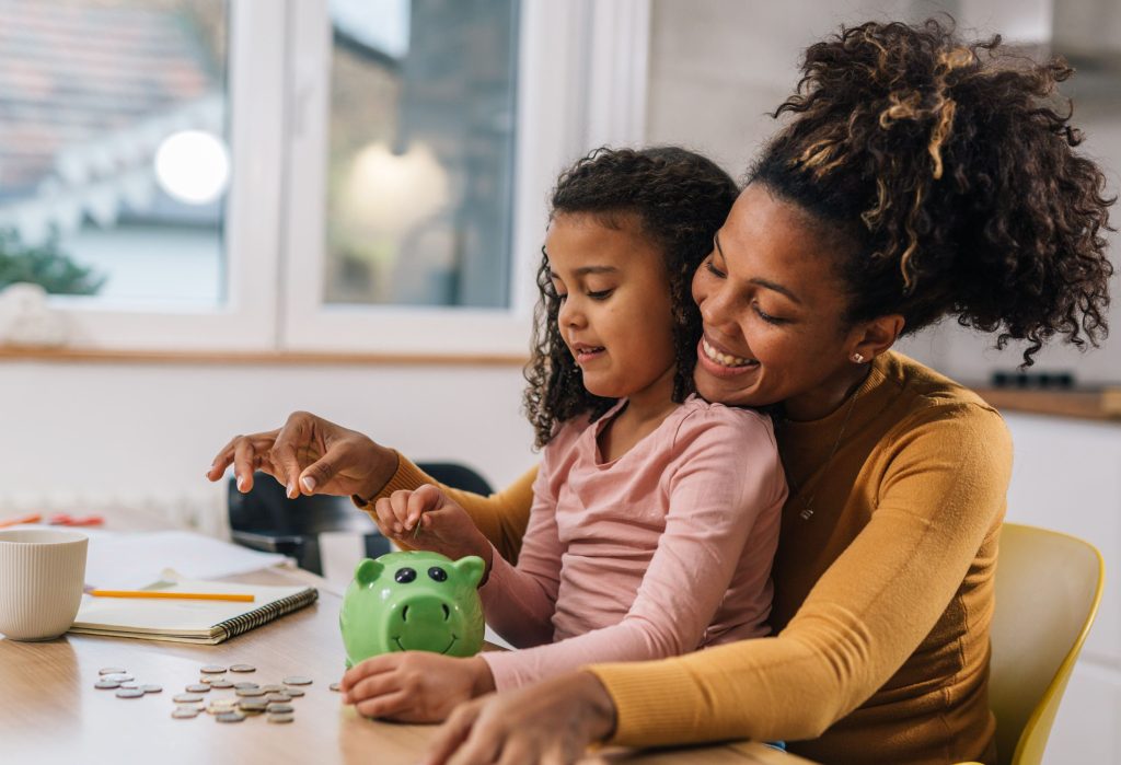 Eine lächelnde Mutter mit hochgestecktem Haar hält ihre kleine Tochter im Arm, während das Kind vorsichtig Münzen in ein grünes Sparschwein auf dem Küchentisch wirft.