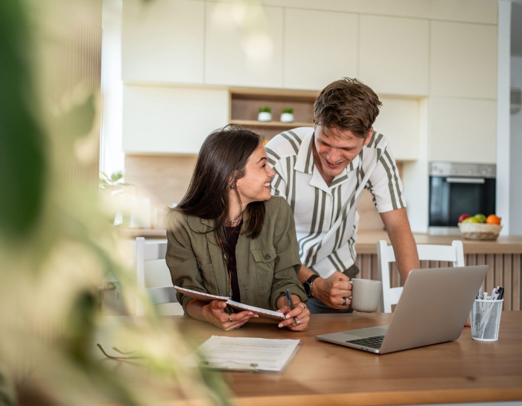 Ein junges Paar interagiert lächelnd an einem Küchentisch aus Holz. Die Frau sitzt und schreibt in ein Notizbuch, während der Mann neben ihr steht, eine Tasse hält und auf den geöffneten Laptop blickt.