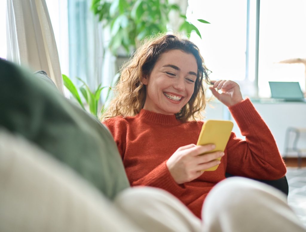 Junge Frau entspannt auf dem Sofa und lacht beim Blick aufs Smartphone – helles, gemütliches Wohnzimmer mit Pflanzen im Hintergrund.