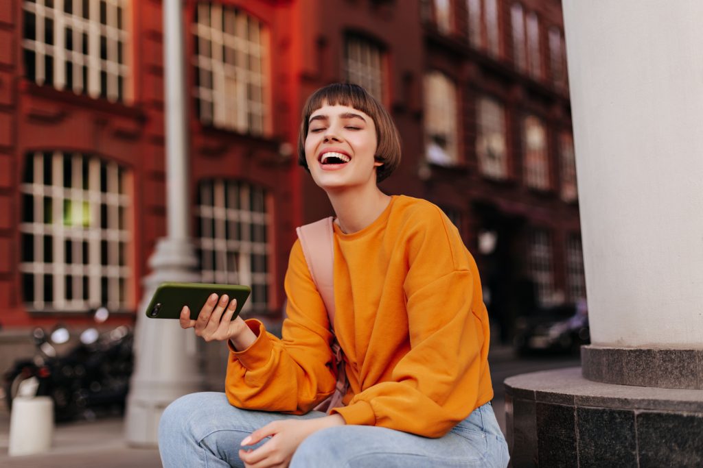 Eine junge Frau mit kurzem braunem Haar und orangefarbenem Pullover sitzt lachend im Freien vor einem roten Backsteingebäude und hält ein Smartphone in der Hand.