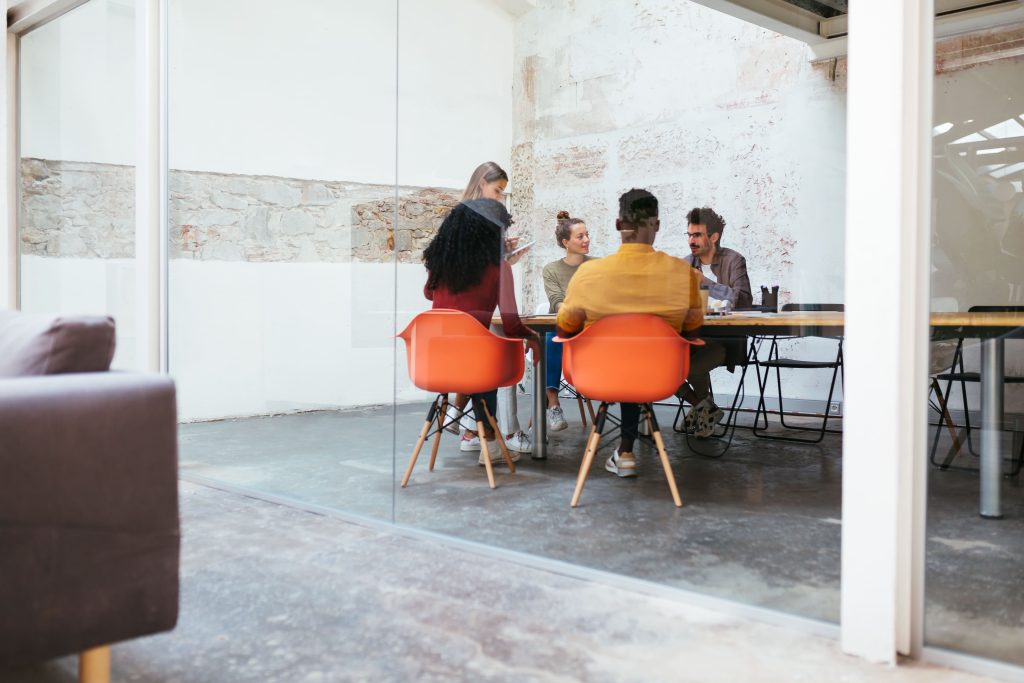 Diverses Team im Meeting hinter Glaswand – moderner Konferenzraum mit Industriecharakter und orangefarbenen Designerstühlen.